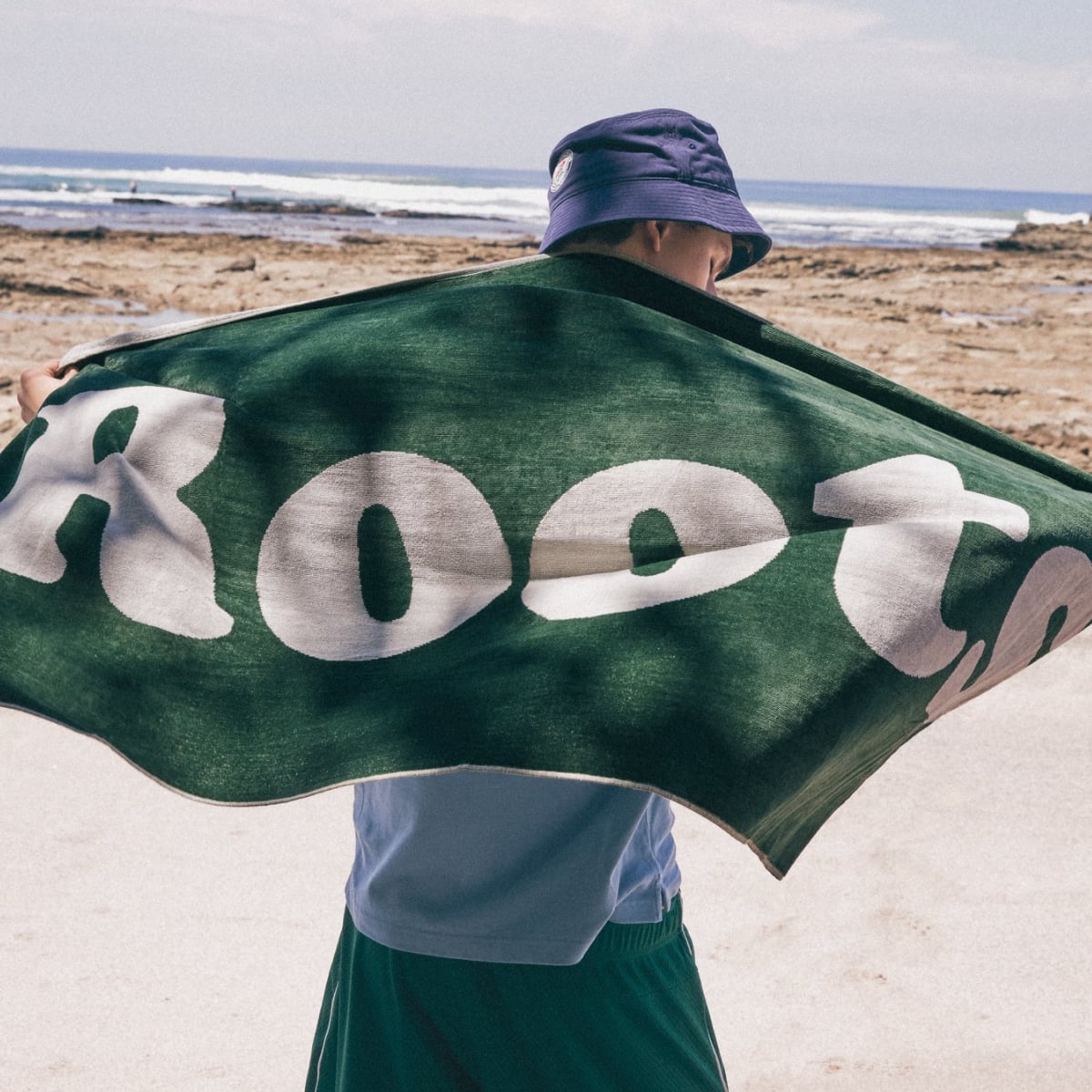 Person on a beach holding up a Roots-branded green towel